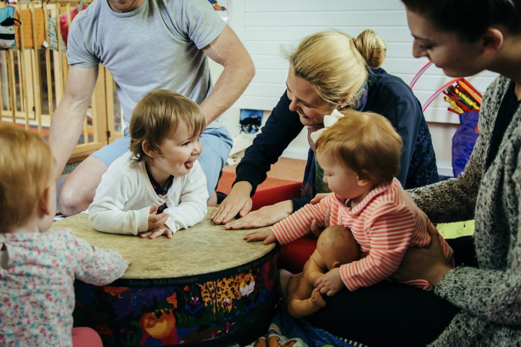 Young children and a woman with blonde hair sit around a large drum and hit it with their hands
