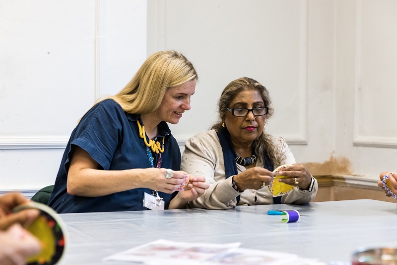 Two women sat at a table sewing. The woman on the left is looking at what the woman on the right is making.