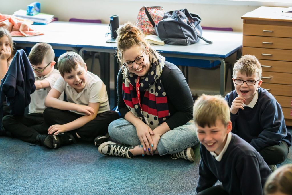 Drama practitioner Emma is sitting cross-legged in jeans and a black top, smiling. She has brown hair in a bun and is wearing glasses. She is surrounded by smiling pupils sitting down cross-legged. 