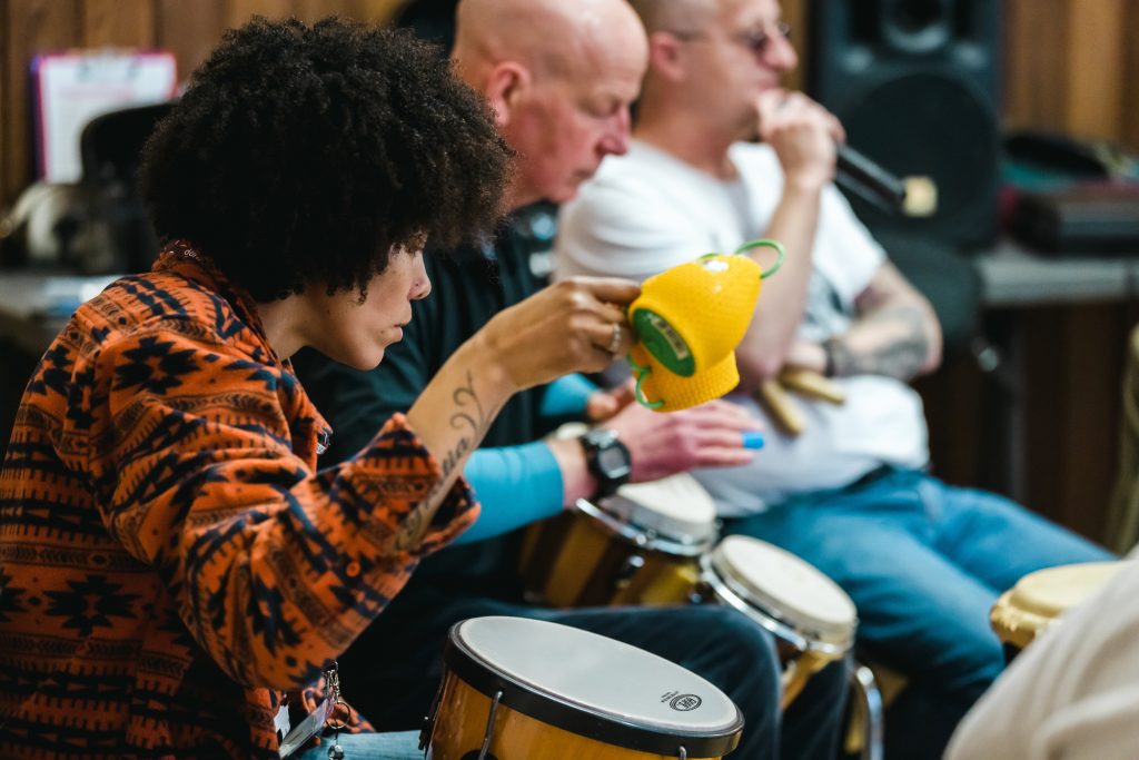 Janet has an afro and wears an orange shirt. She plays a djembe next to two participants 