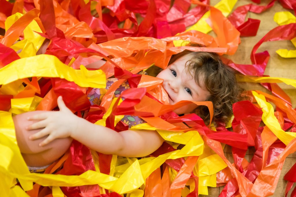 A toddler lies smiling amongst a pile of orange, yellow and red streamers. They are taking part in Tuneful Chatter - a programme for 0-5s to improve personal, social and emotional development through creativity. A Tuneful Chatter session led by darts in Doncaster at The Point centre for creative health.