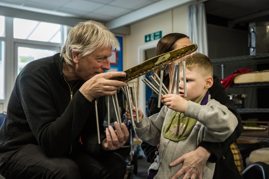 An adult with short-ish grey hair and dark clothing sits and holds a musical instrument with lots of metal rods hanging down. A child with short fair hair and a grey sweatshirt is playing with the instrument. The adult is looking at the child and the child is looking at the instrument, they are both in a classroom. This is part of All Aboard, a project in Doncaster led by darts