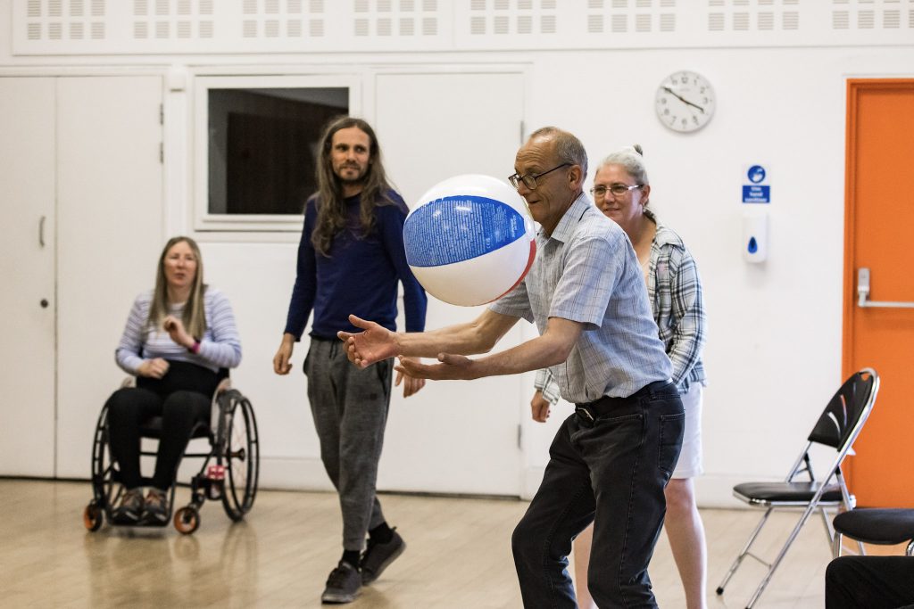 our adults are taking part in a Creative Directions Moves session run by darts in Doncaster. Three adults are standing and one is seated in a wheelchair. One of the standing adults is holding their arms out in front of them, about to catch and inflatable beach ball.