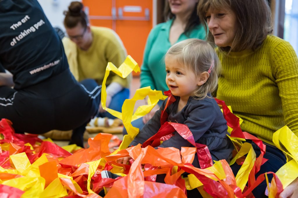 A young child sits in a pile of red and yellow streamers, smiling, whilst their adult sits behind them at a Tuneful Chatter session. This is a darts in Doncaster session at The Point