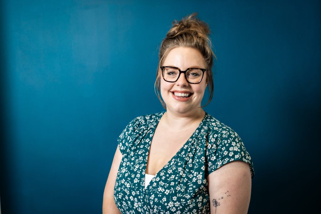 Emma Liversage, a drama practitioner, has long hair in a bun on her head, glasses and a big smile. She we are a green flowery dress.