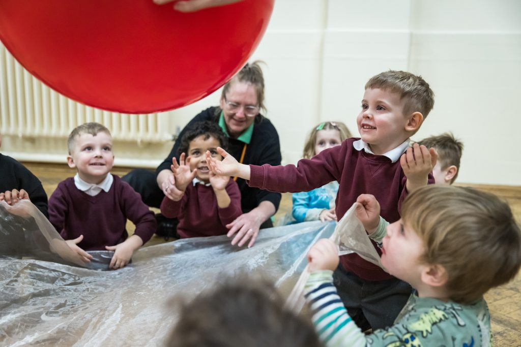 A group of young children and nursery staff sit on the floor, laughing and playing with a large red ball during a Tuneful Chatter session at a nursery in Doncaster