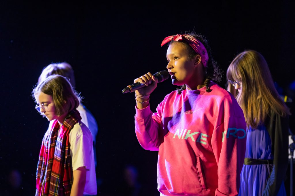 A group of young people wearing bright colours sing into microphones on stage at The Dome in Doncaster - part of iSing for Doncaster Music Hub led by darts