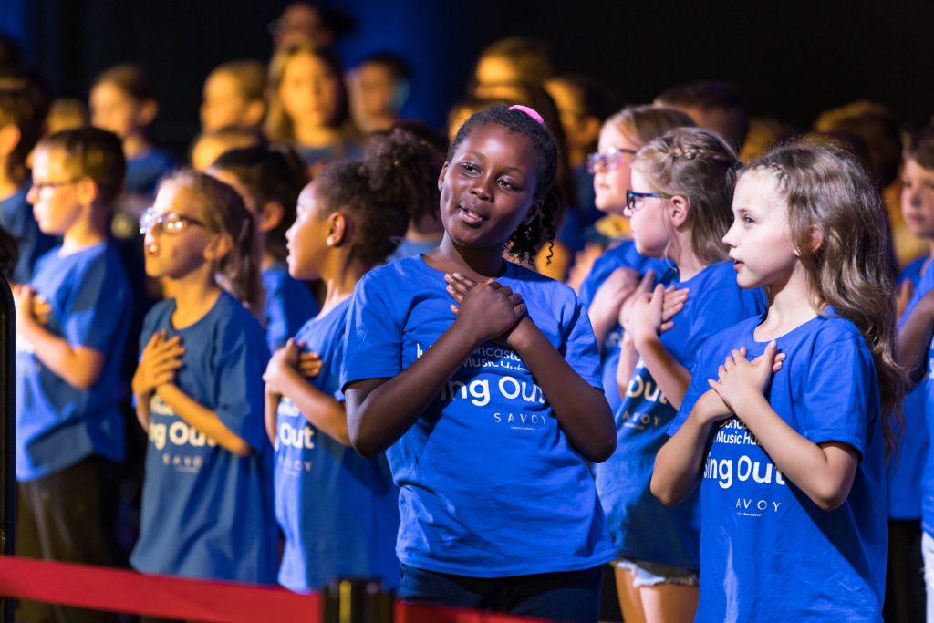 A group of young people in bright purple Sing Out t-shirts smile and sing at last year's Sing Out event at The Dome. This is a Doncaster Music Hub event led by darts