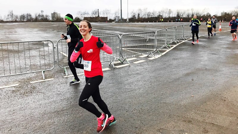 An individual is running in an organised race. They are wearing a pink top, black leggings, pink trainers and black gloves. They have a big smile on their face, and they have their thumbs up.