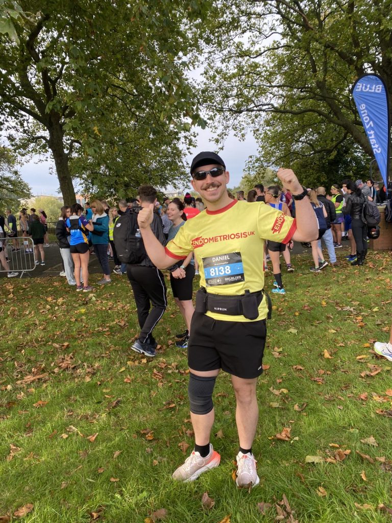 An individual, in cap and sunglasses, is smiling and has just ran a race. They wear a yellow t-shirt, black shorts and white trainers. They have their thumbs up, celebrating, and are surrounded by grass and woodland.
