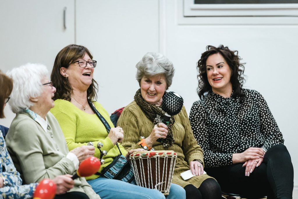 Four women laugh and singing, playing the bongos and maracas in a group by darts.