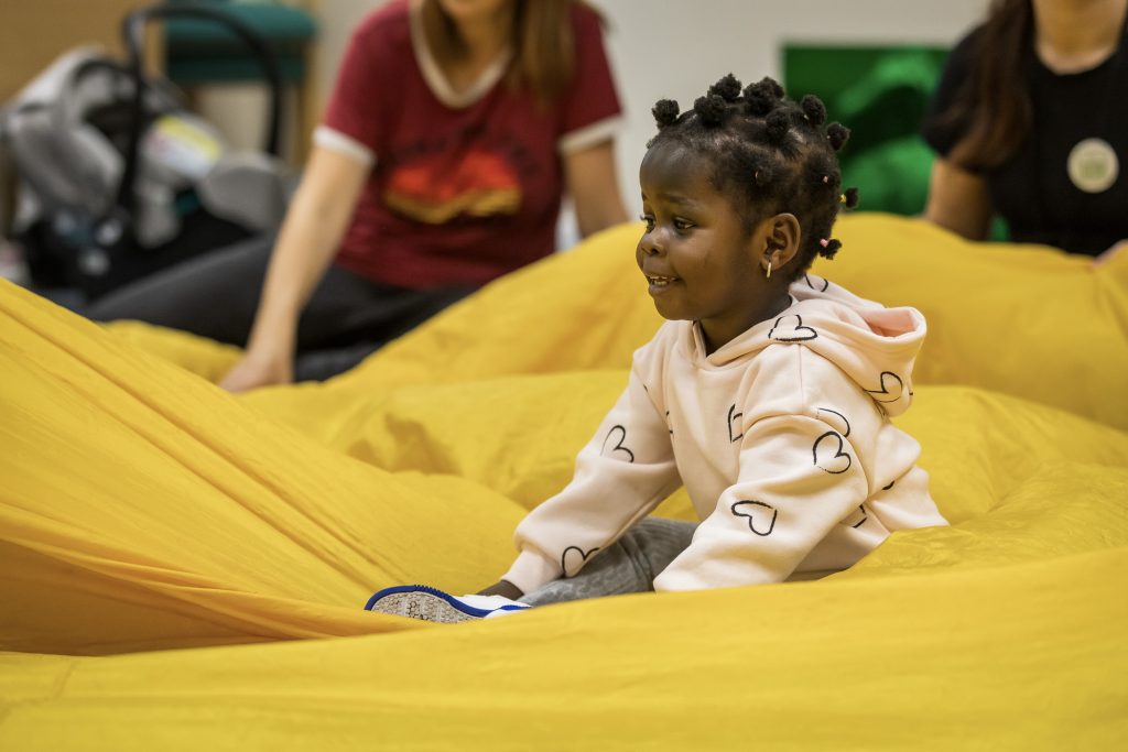 A young child wearing a peach coloured hoodie sits on a yellow parachute, as the parachute is waved around them by adults and children sitting around the edge.