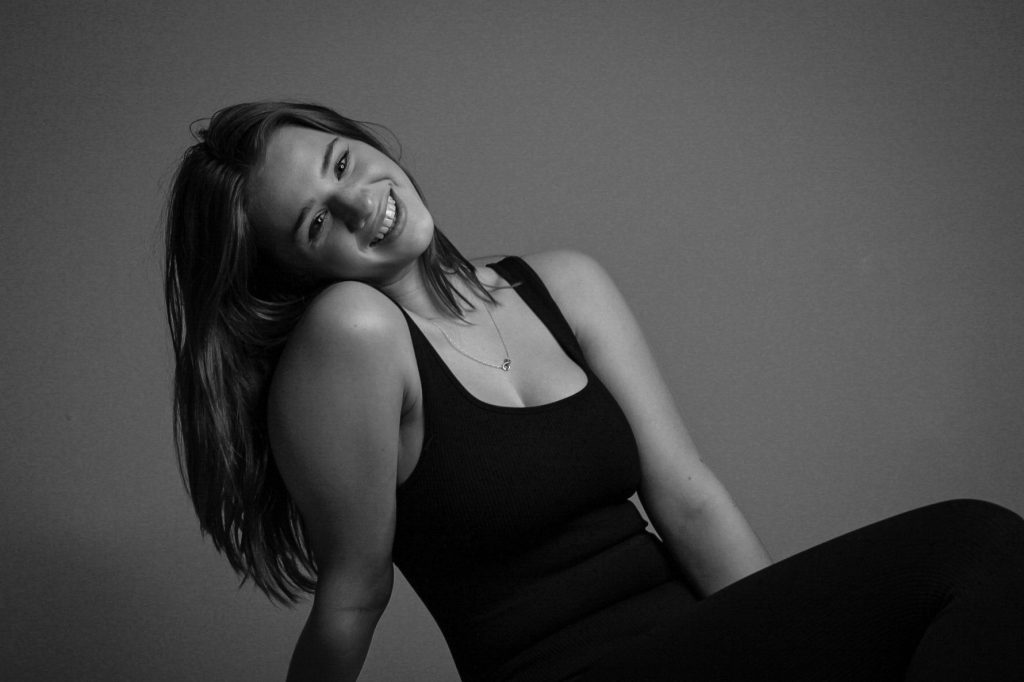 A young woman smiles at the camera, sat on the floor. She has long dark hair and wears a black vest and leggings. The image is in black and white. 