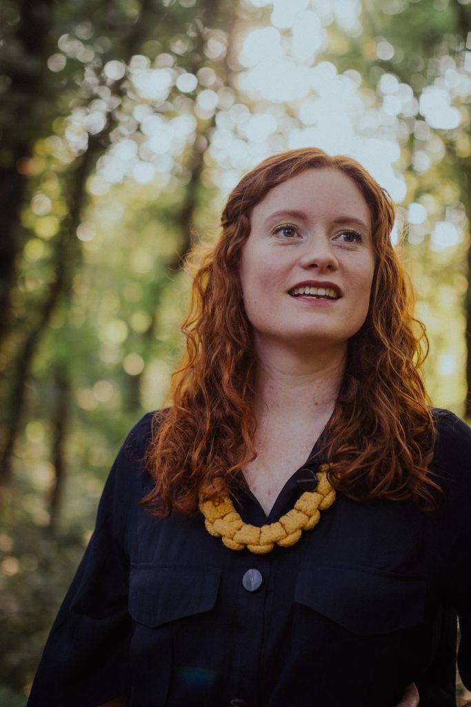 A lady with red hair looks away from the camera. She is in a forest and surrounded by trees.