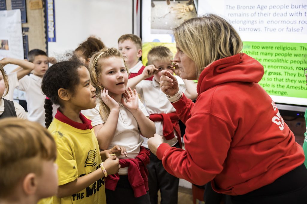 A group of children wearing red and white school uniforms stand in front of an adult who is bending down to talk to them, gesturing with their hands as they explain an activity.