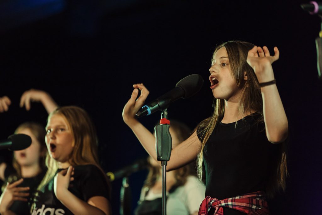 Three girls sing on stage into a microphone, with their hands up in action. They are part of darts' Swag Choir. 