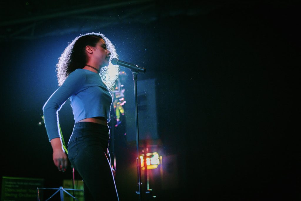 A girl sings into a microphone. She has curly afro hair and wears a blue top. She is part of darts' Swag Choir. 