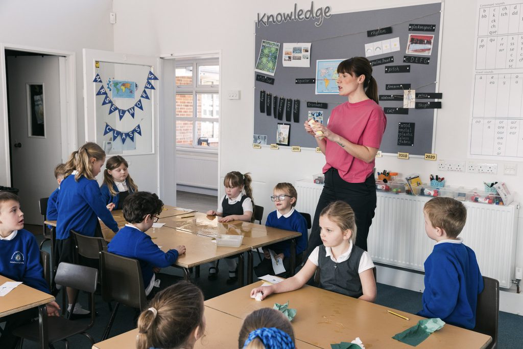A teacher with brown hair, a pink top and black trousers stands up, teaching a classroom of young children. She is part of a darts programme for teachers.