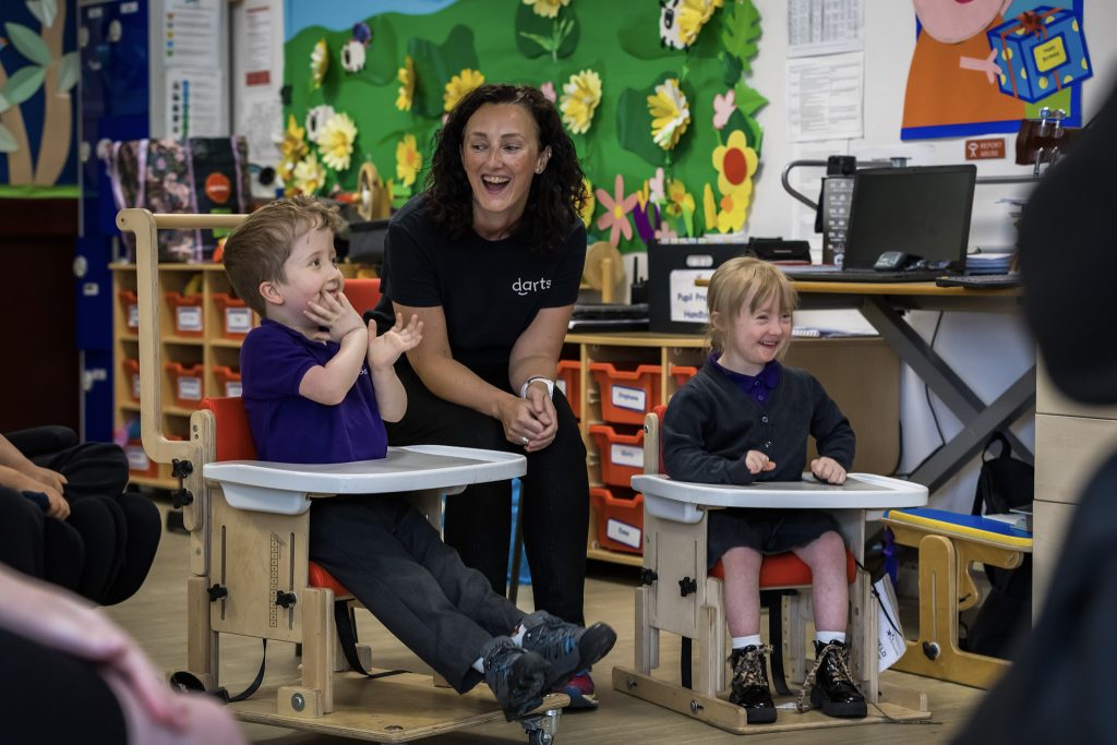 A classroom scene features a darts artist and two smiling young children in specialised adaptive seating. The artist, with dark, curly hair and wearing a black 