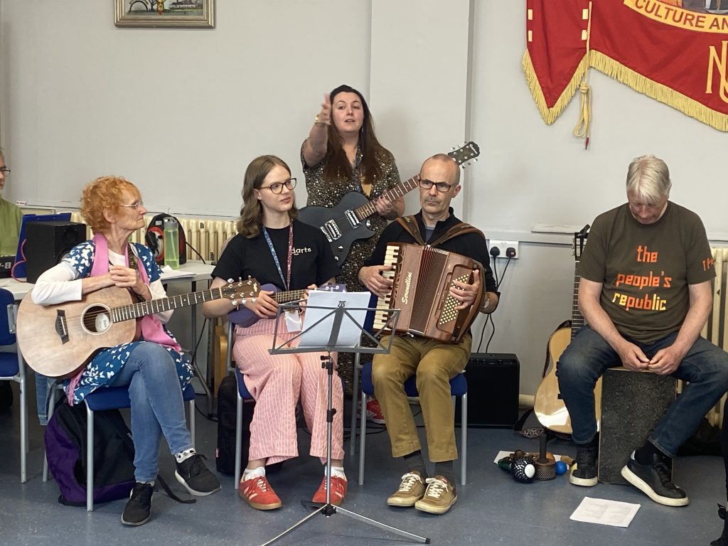 A group of five musicians of varying ages and genders sit in a semi-circle, playing instruments, including an acoustic guitar, ukulele, electric guitar, accordion and Cajon percussion box.