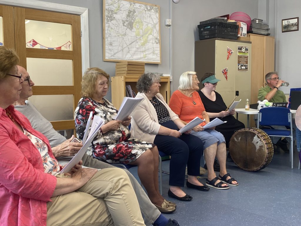 A group of adults sitting in chairs at a celebration event in a community centre. Most are holding song sheets in their hands and singing.
