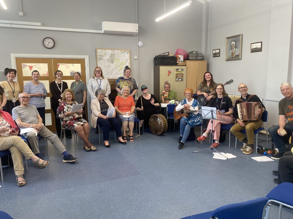 A large group of people of mixed ages and genders are sitting in a semi-circle in a brightly lit room in a community centre. Some are holding musical instruments, including a guitar, ukulele, accordion, and drum. Several others hold sheets of music.