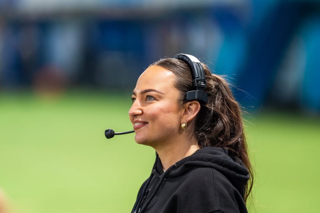 A young woman with brown hair in a ponytail and on overhead mic stands on a sports field, looking up in awe. 