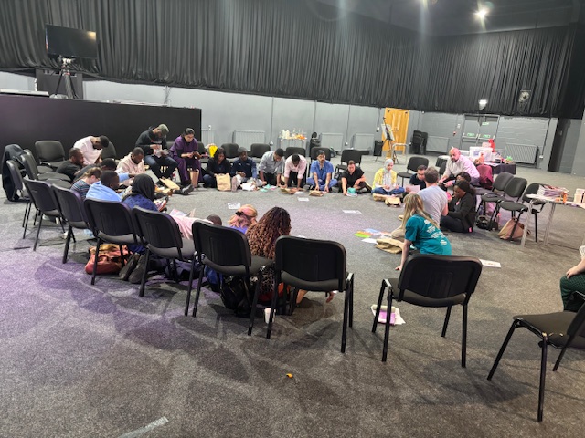 A group of approximately 25 diverse people sit cross-legged on the floor, forming a circle inside of a large grey carpeted room with a dark curtained backdrop. 