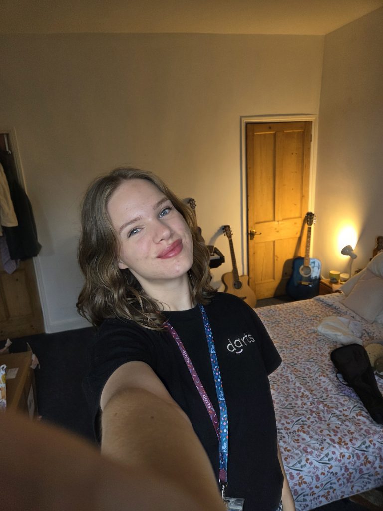 A young woman takes a selfie - smiling at the camera. She wears a darts branded t-shirt and lanyard, and has short brown hair in a bob. 