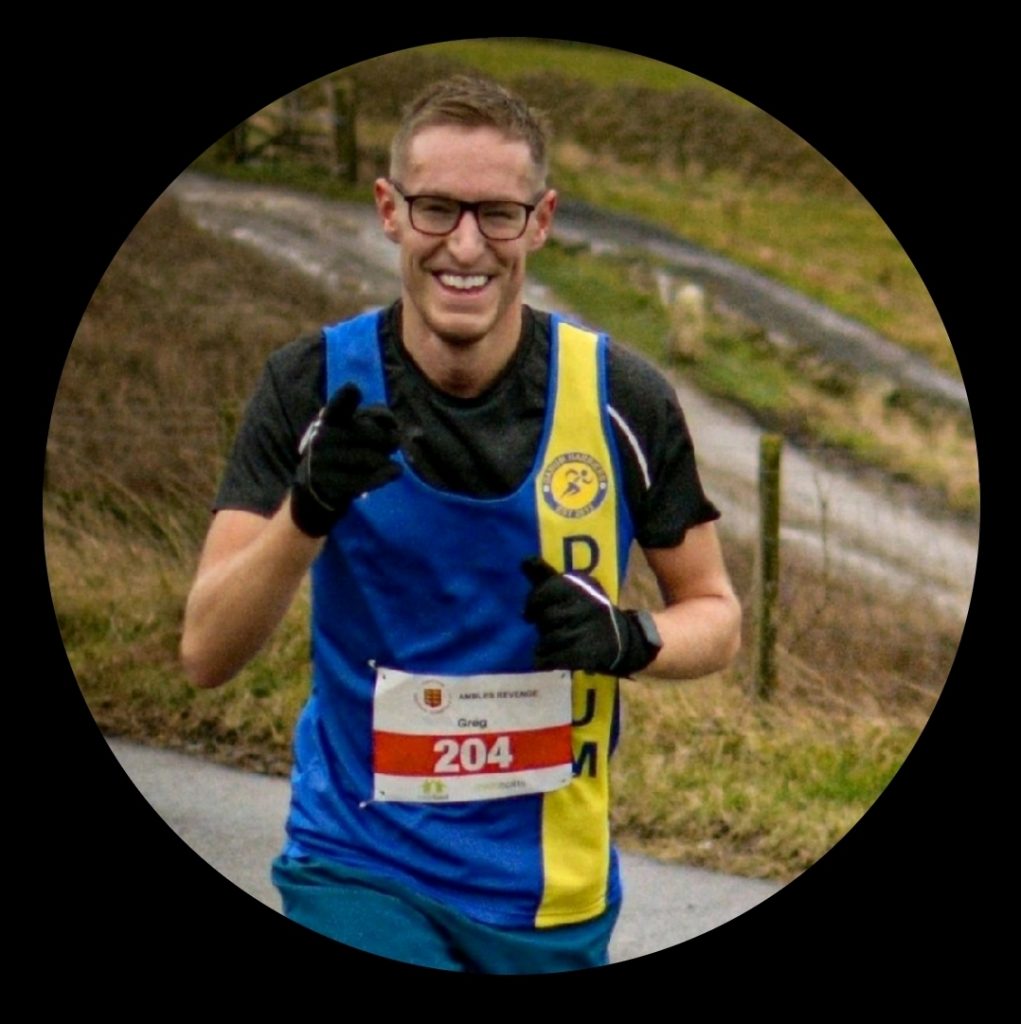 A man runs in an organised race. He has short brown hair and glasses. He smiles widely at the camera and points. 
