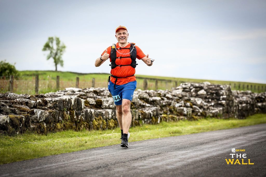 A man runs along a country lane in a red top, red cap, blue shorts and black trainers. He is smiling widely at the camera - with his thumbs up. 