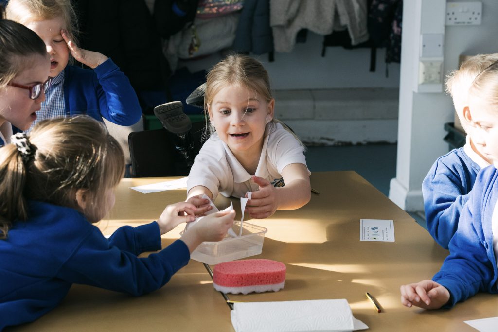 A child wearing a white tshirt leans over a wooden table, reaching for items in the middle, with an excited expression on their face, during a creative session run by darts in Doncaster