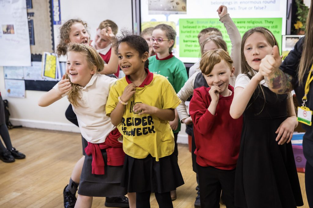 A group of children wearing red and white school uniforms stand in a group and giggle during a performance of their work, as part of a creative session led by darts in Doncaster.