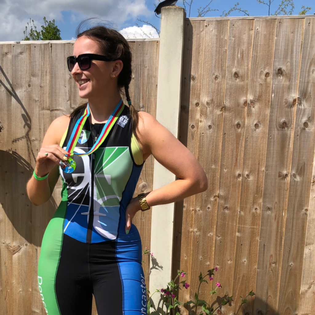 A woman with brown hair in two French plaits smiles while holding her Triathlon medal. She wears Triathlon gear and sunglasses and is stood outside. 
