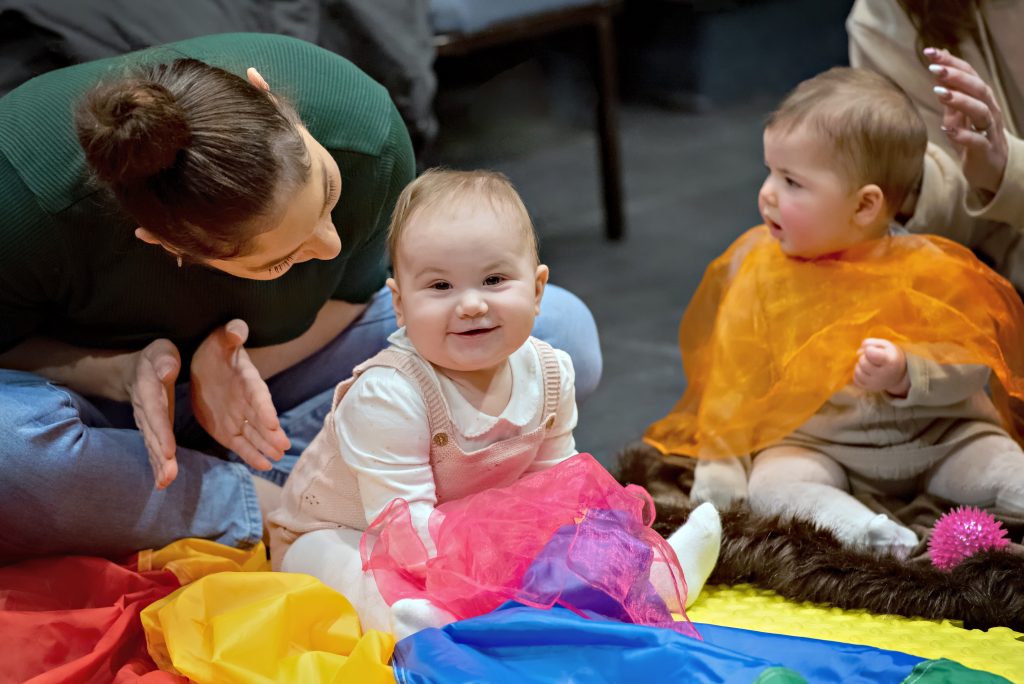 Two babies sit on the floor and play with colourful fabrics in a Hello Baby session at Cast in Doncaster.