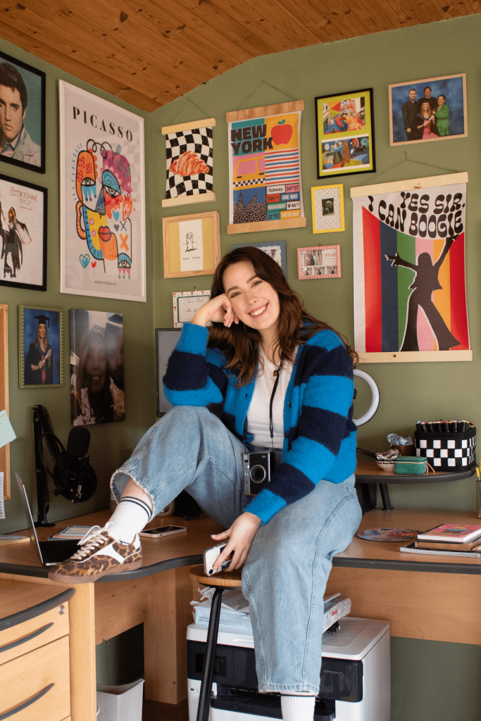 A woman with long brown hair wearing a striped cardigan, jeans and trainers sits on her desk and smiles at the camera. She is surrounded by prints and art work.