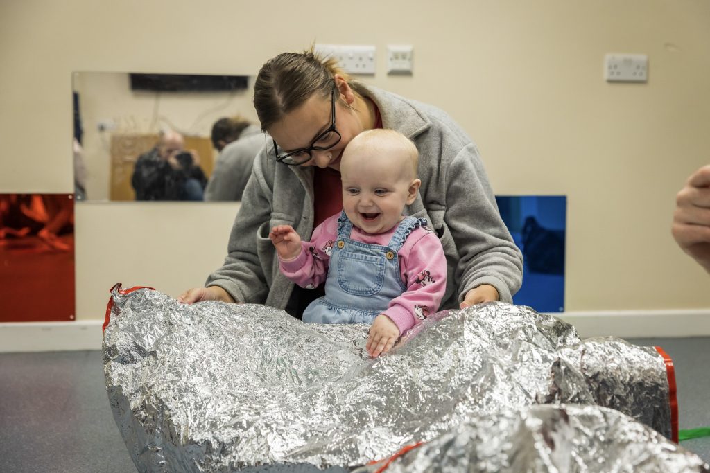 A baby sits on their adult's lap and plays with a foil blanket, laughing and scrunching it in their hands during a Tuneful Chatter session from darts in Doncaster