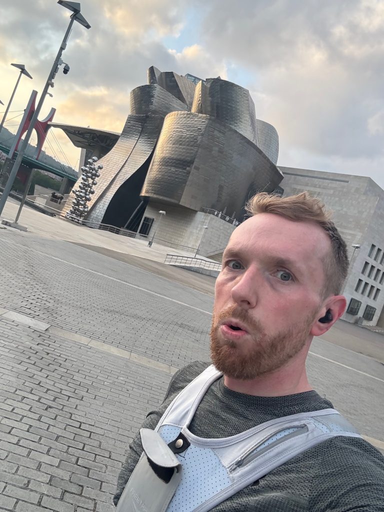 A man looks into the camera whilst on a run. There is a scenic city background behind him. 