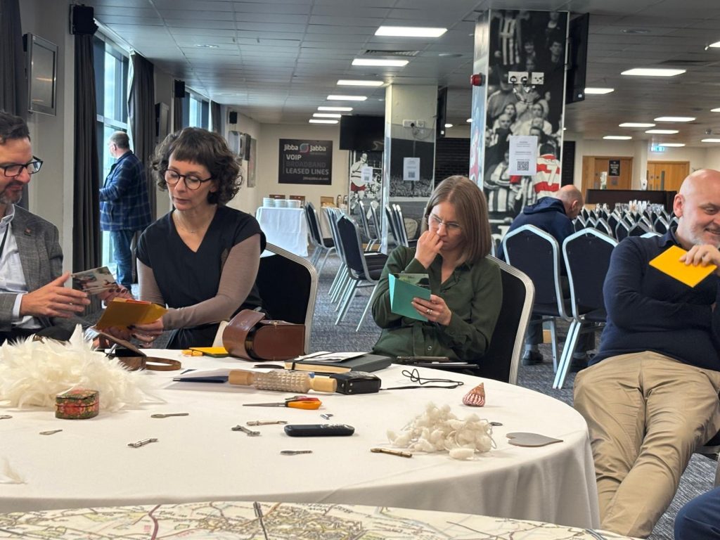 A group of adults participating in a darts workshop at the Team Doncaster Partnership Summit 2025. They are seated around a round table covered with a white tablecloth, on which various vintage tools and random objects are scattered. Another table, partially visible in the foreground, is covered with a map tablecloth.
