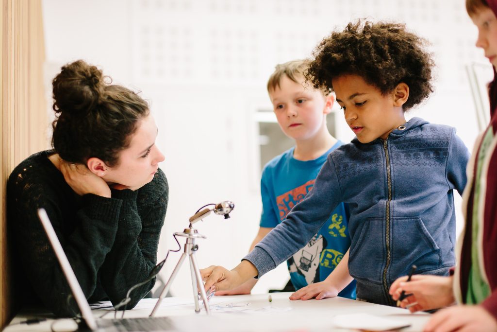 A young adult leans forward on a table wearing a black jumper. They have long dark hair up in a bun. Two children are using animation equipment and standing next to the table.