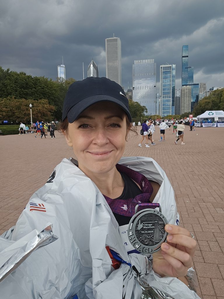 A woman smiles into the camera. She is wearing a cap and a foil blanket, holding a medal. She has just completed a marathon. 
