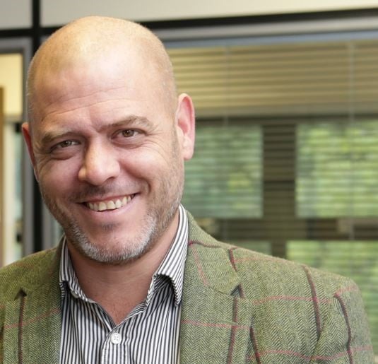 A man smiles at the camera. He is bald and wearing a tweed jacket. He is taking part in dash for darts.