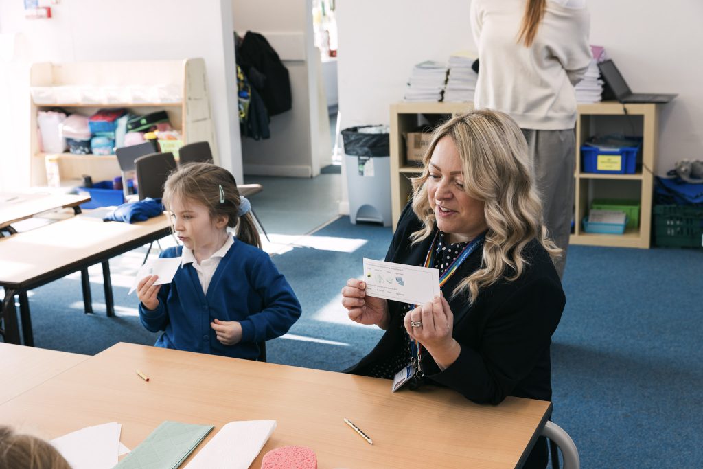 A teacher and young pupil are sat side by side in a classroom. They are expressive and are working with flashcards.
