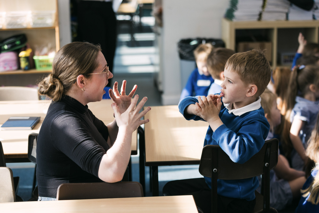 A teacher works with a young pupil, demonstrating hand gestures. They are sitting opposite each other in a classroom.