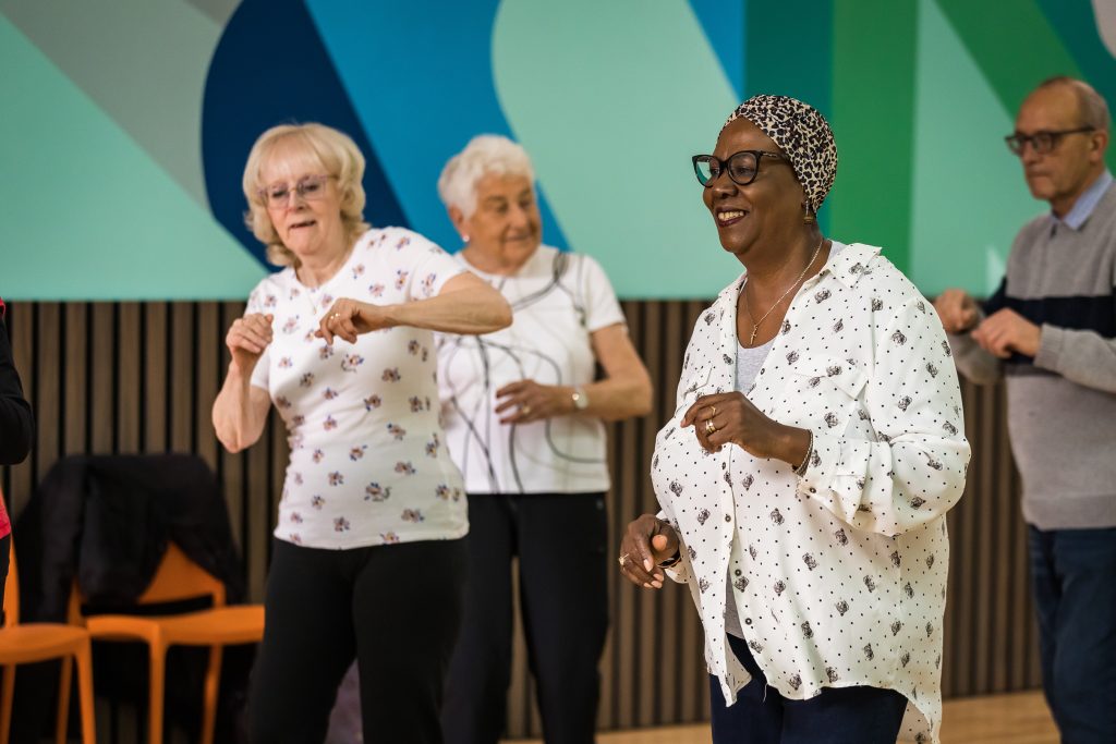 A group of adults dance and smile against a colourful painted background. A Dance On session led by darts at The Point in Doncaster