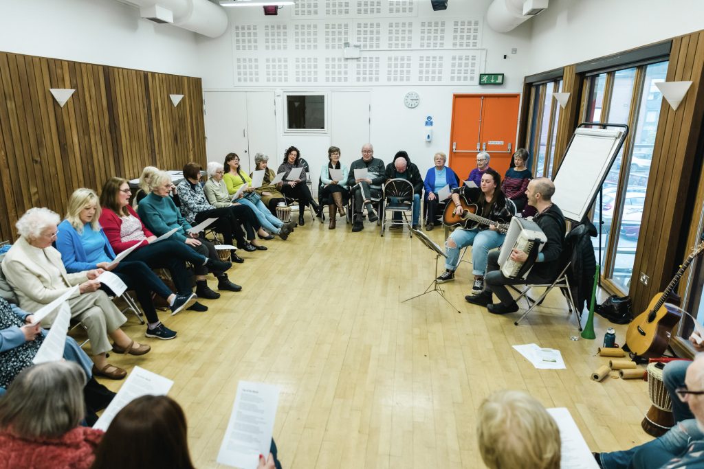 A group of adults are seated in a circle, singing and holding song lyric sheets. Part of a Singing for Memory session run by darts at The Point in Doncaster.
