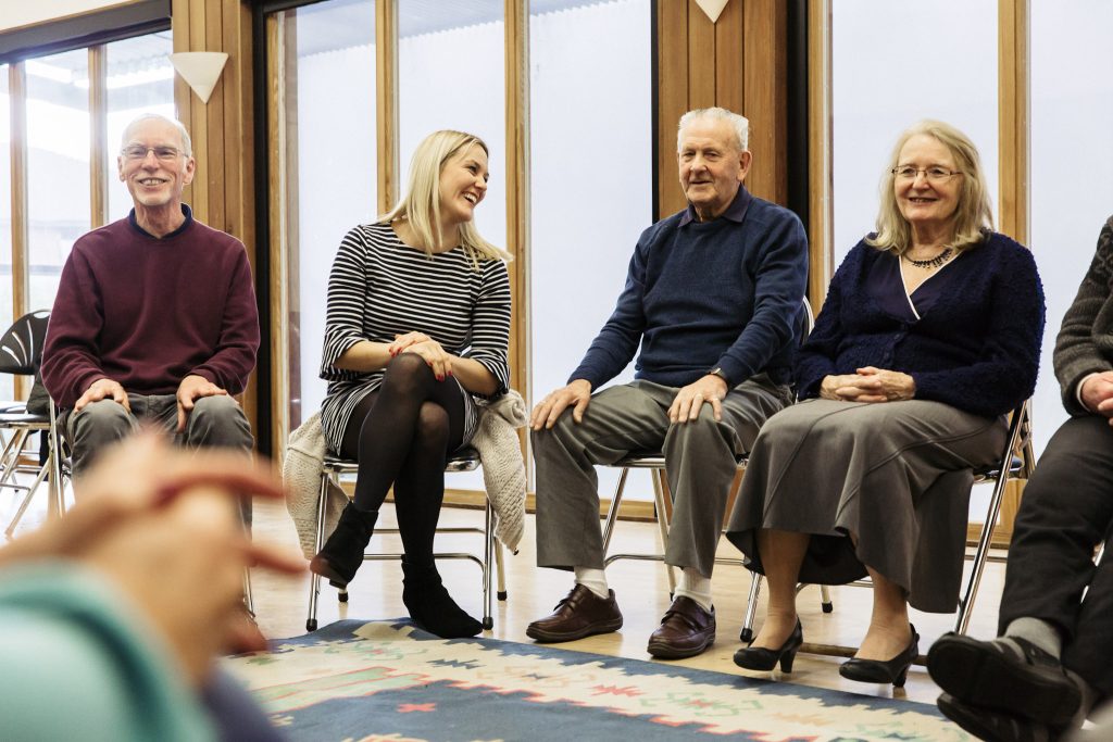 A diverse group of adults sitting in a circle are singing in a brightly lit room with large windows. Part of a Singing for Memory session run by darts at The Point in Doncaster.