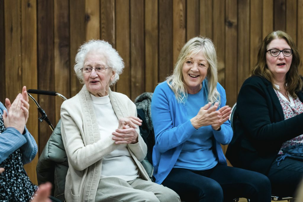 Three women seated, singing and clapping, with wooden panelling in the background. Part of a Singing for Memory session at The Point, home of darts, in Doncaster.