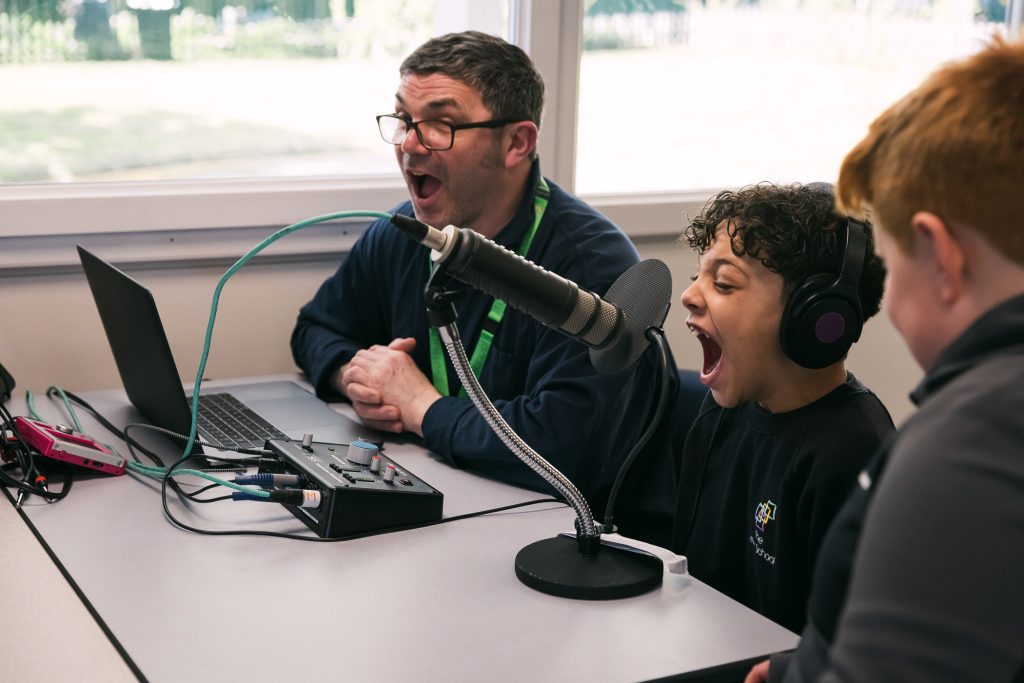 Artist Moony and a young person smile and sing with their mouths wide open. The young person has headphones on and a microphone. This is a darts project in Doncaster