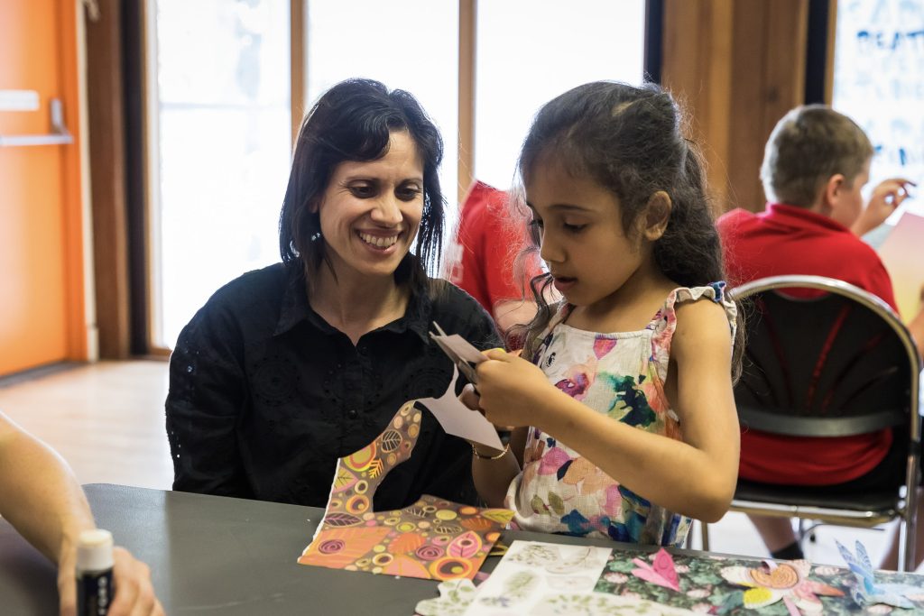 An adult and child are seated next to each other at a table. The adult watches and smiles as the child carefully cuts out shapes to add to their art work.